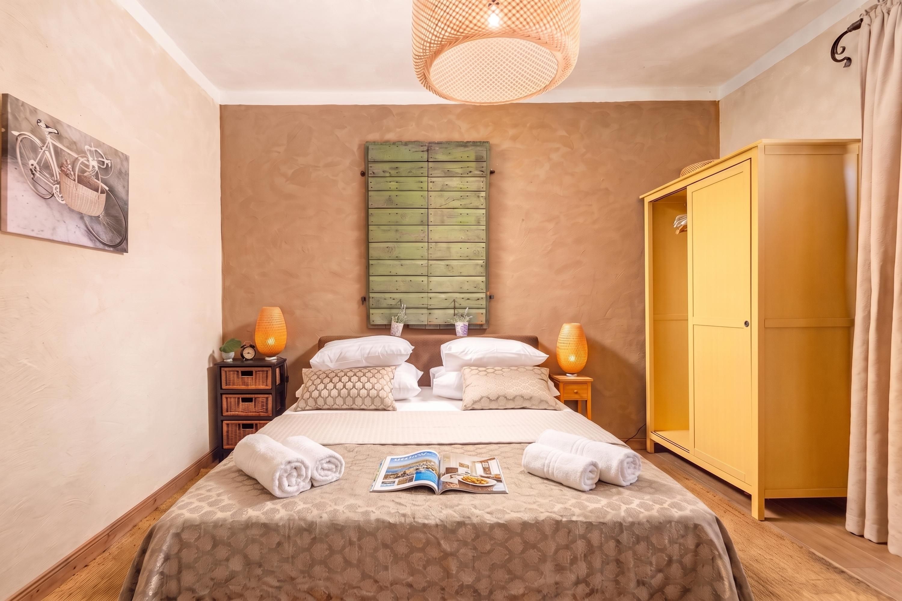 A neatly arranged bedroom with a bed, a wardrobe, a bedside table with a lamp, and a wall-mounted shelf.