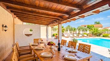 An outdoor dining area with a pool view, wooden tables, and chairs.