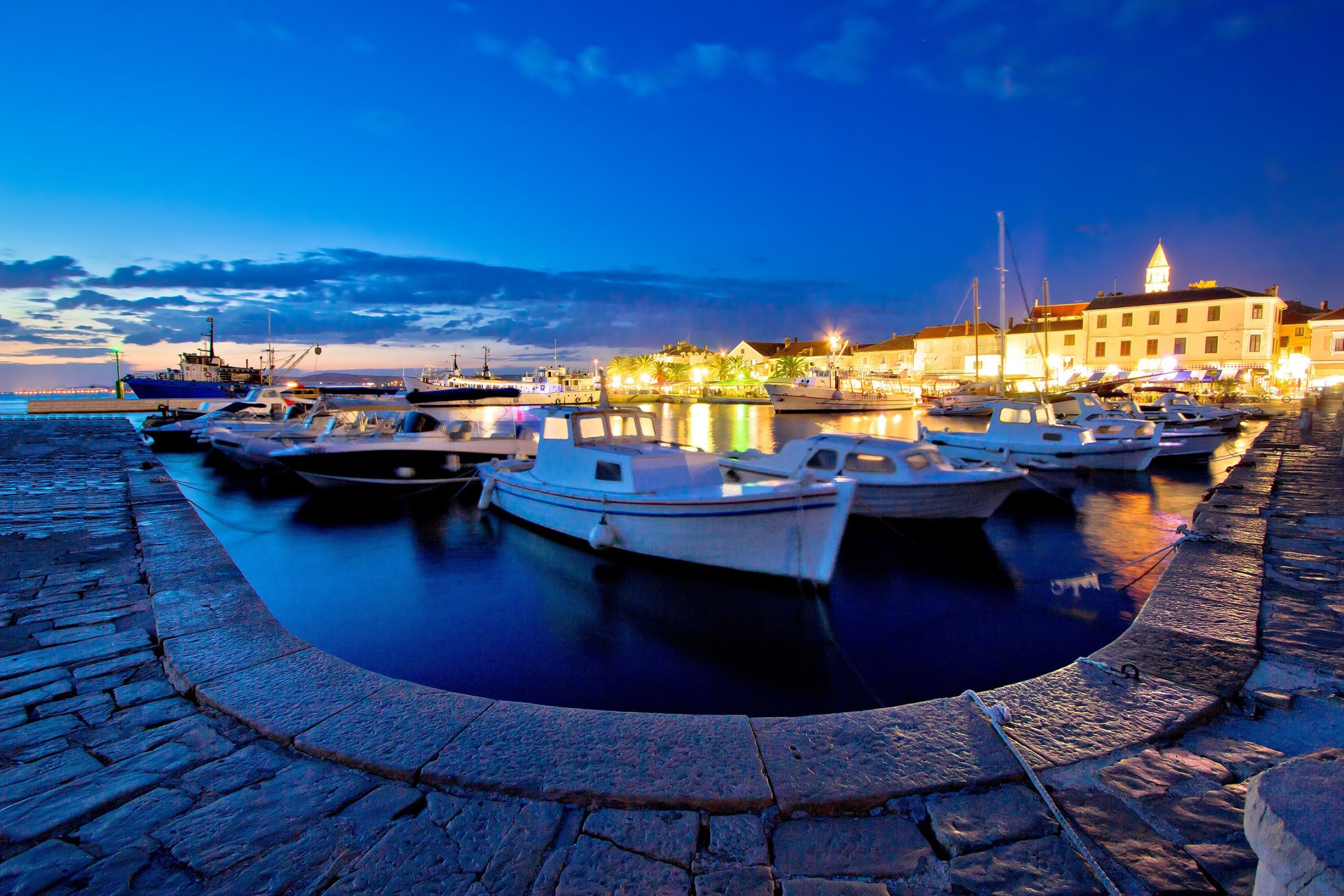 A marina with boats docked, a stone-paved waterfront, and buildings in the background.