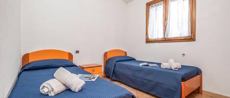 A room with two beds, each with a blue bedspread and an orange headboard. There is a window with wooden frames and white curtains.