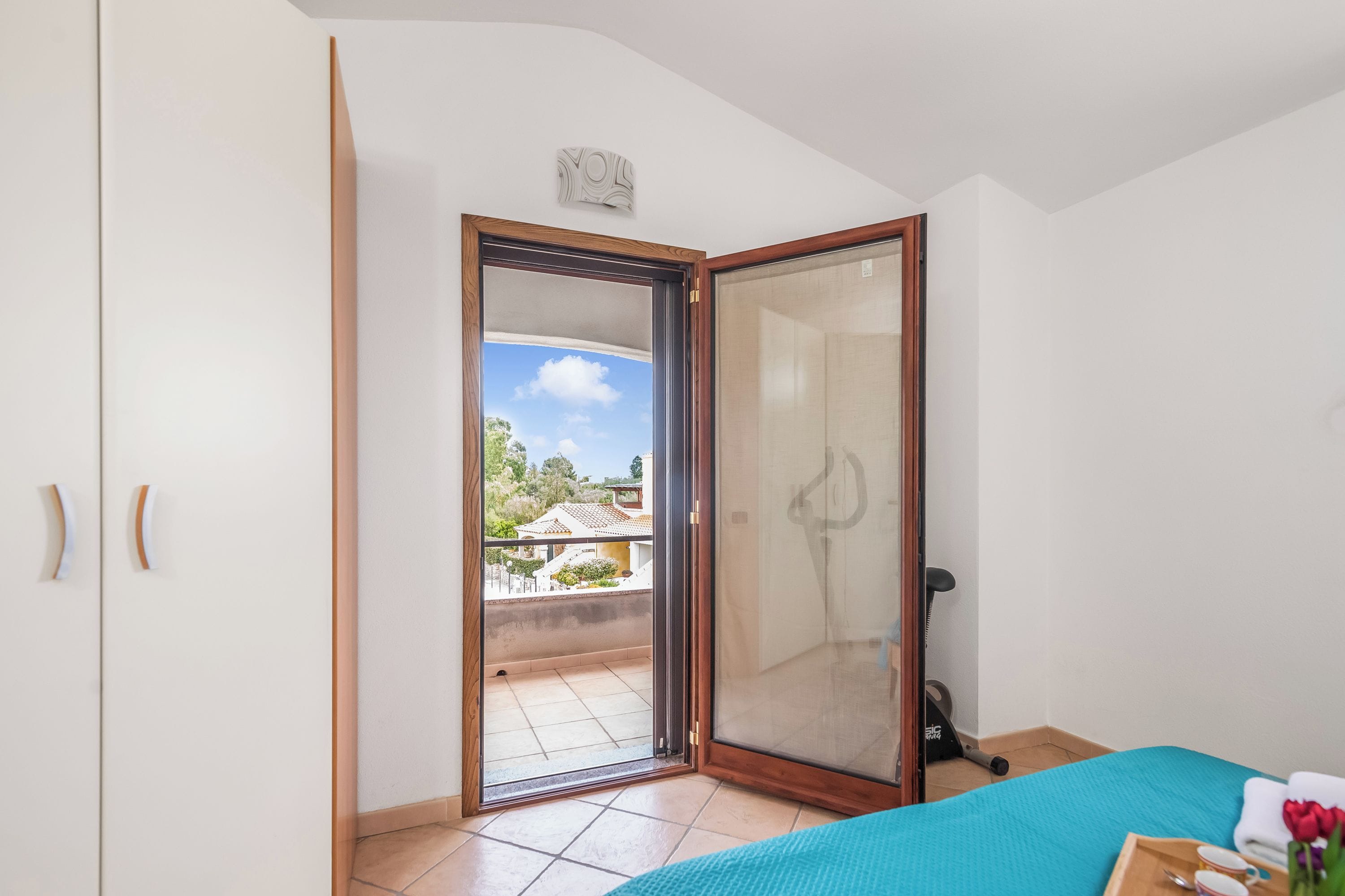 Une pièce dotée d’une porte-fenêtre coulissante donnant sur un balcon, d’un lit recouvert d’une couverture bleue et d’une petite table sur laquelle sont posés divers objets.