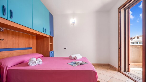 A hotel room with a bed, a wooden wardrobe, a small table, and a view of the outside. - Budoni Beach Homes (Budoni)