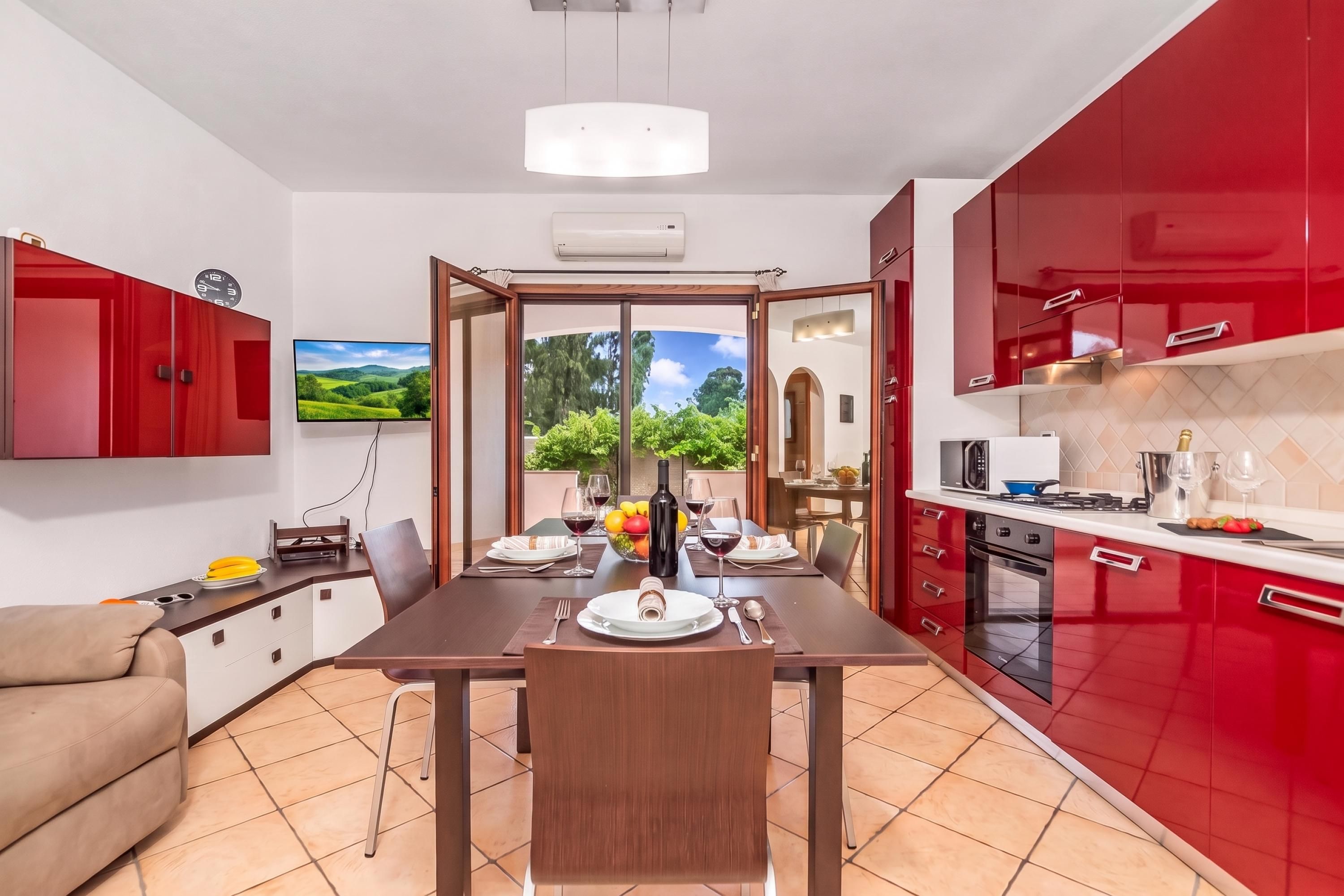 A modern kitchen with red cabinets, a dining table set for two, and a view of greenery outside.