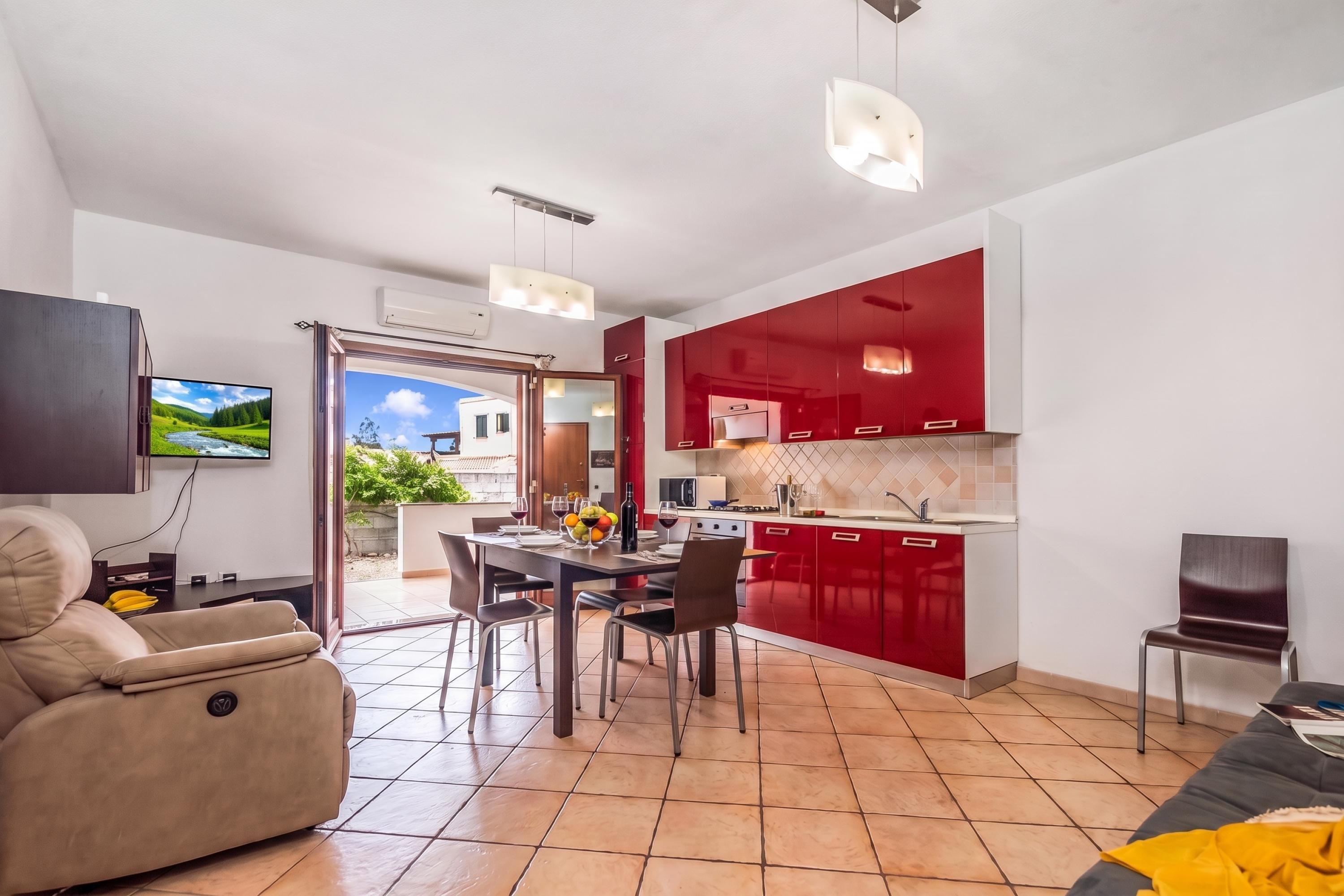 A modern living room with a sofa, a dining table set for four, and a kitchen with red cabinets.