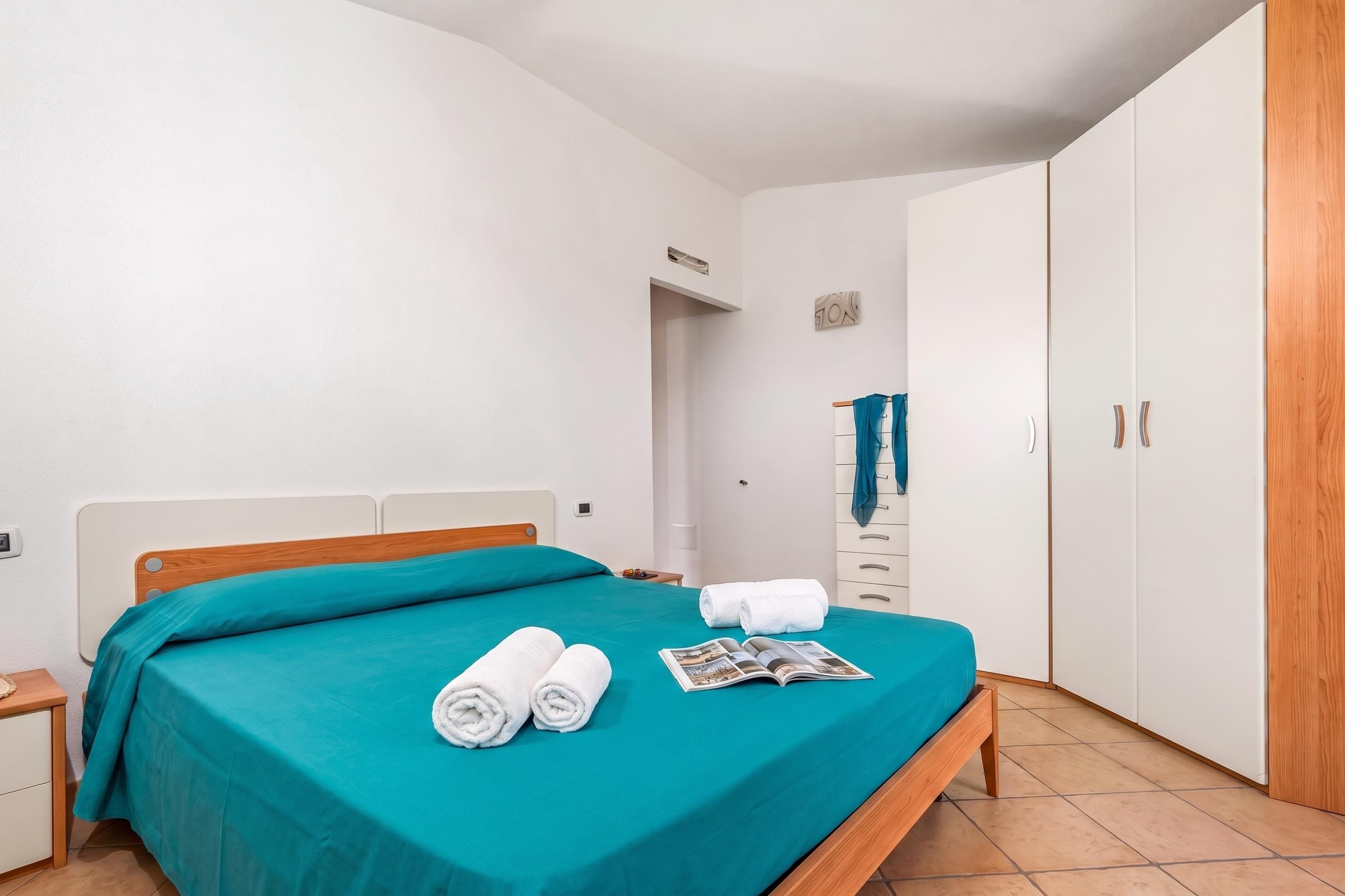 A neatly made bed with a teal bedspread, two rolled white towels, and a magazine on the bed.