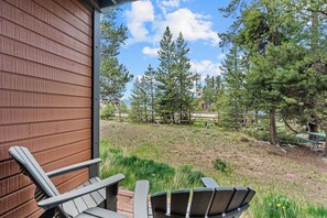 A wooden exterior wall, a wooden deck with two Adirondack chairs, and a view of a grassy area with trees and a bench.