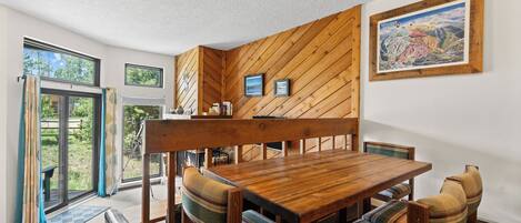 Un espace repas comprenant une table et des chaises en bois, un banc et une vue sur l’extérieur, accessible par des portes vitrées.