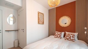 Un dormitorio con cama, mesita de noche, espejo, cuadro enmarco y lámpara de pared.