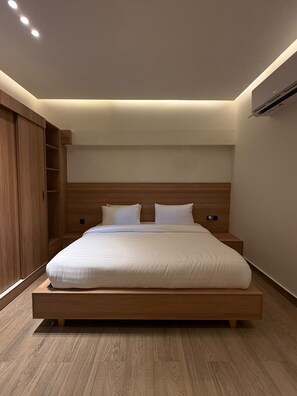 A modern bedroom with a wooden bed frame, white bedding, and built-in shelving. - Hamsun Dha Modern Family Suite Karachi (Karachi)