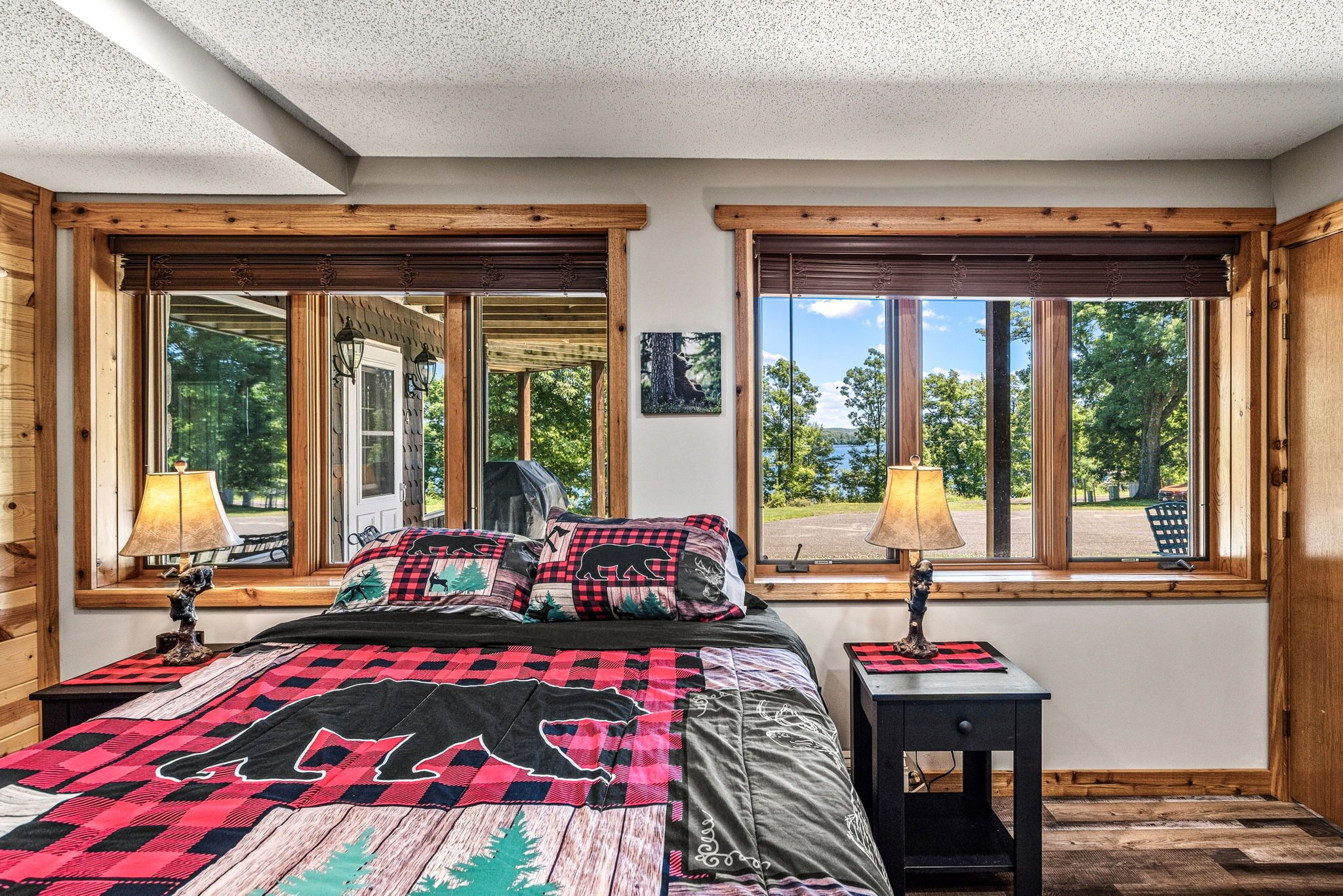 A bedroom with a large bed, two windows, a nightstand, and a lamp.