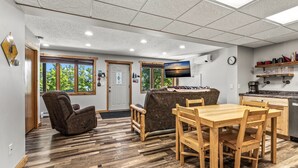 Una habitación con una mesa de comedor de madera y sillas, un sillón de masaje, un sofá, un televisor y una zona de cocina con estanterías.