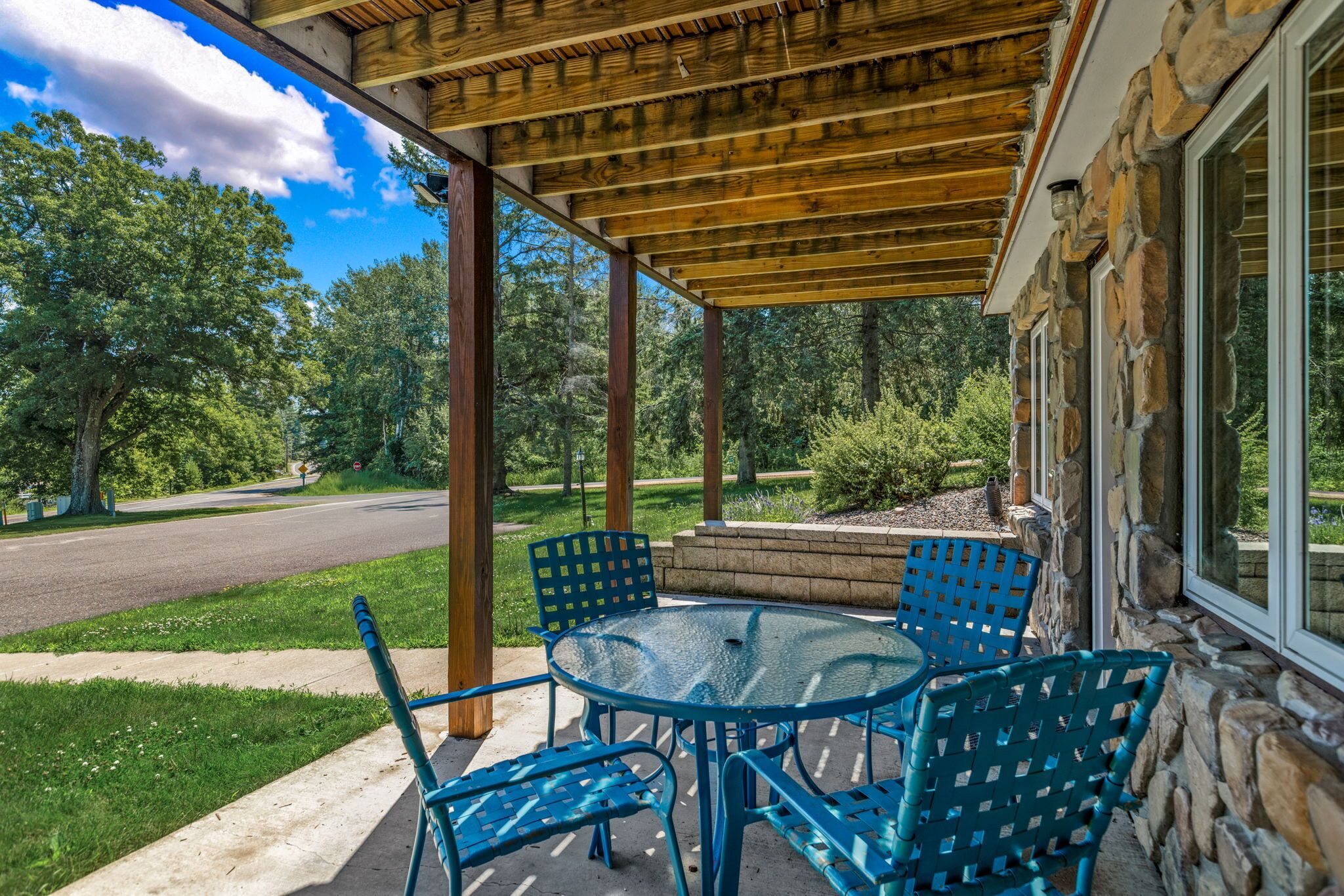 A covered patio area with a stone wall, a glass table, and blue chairs.