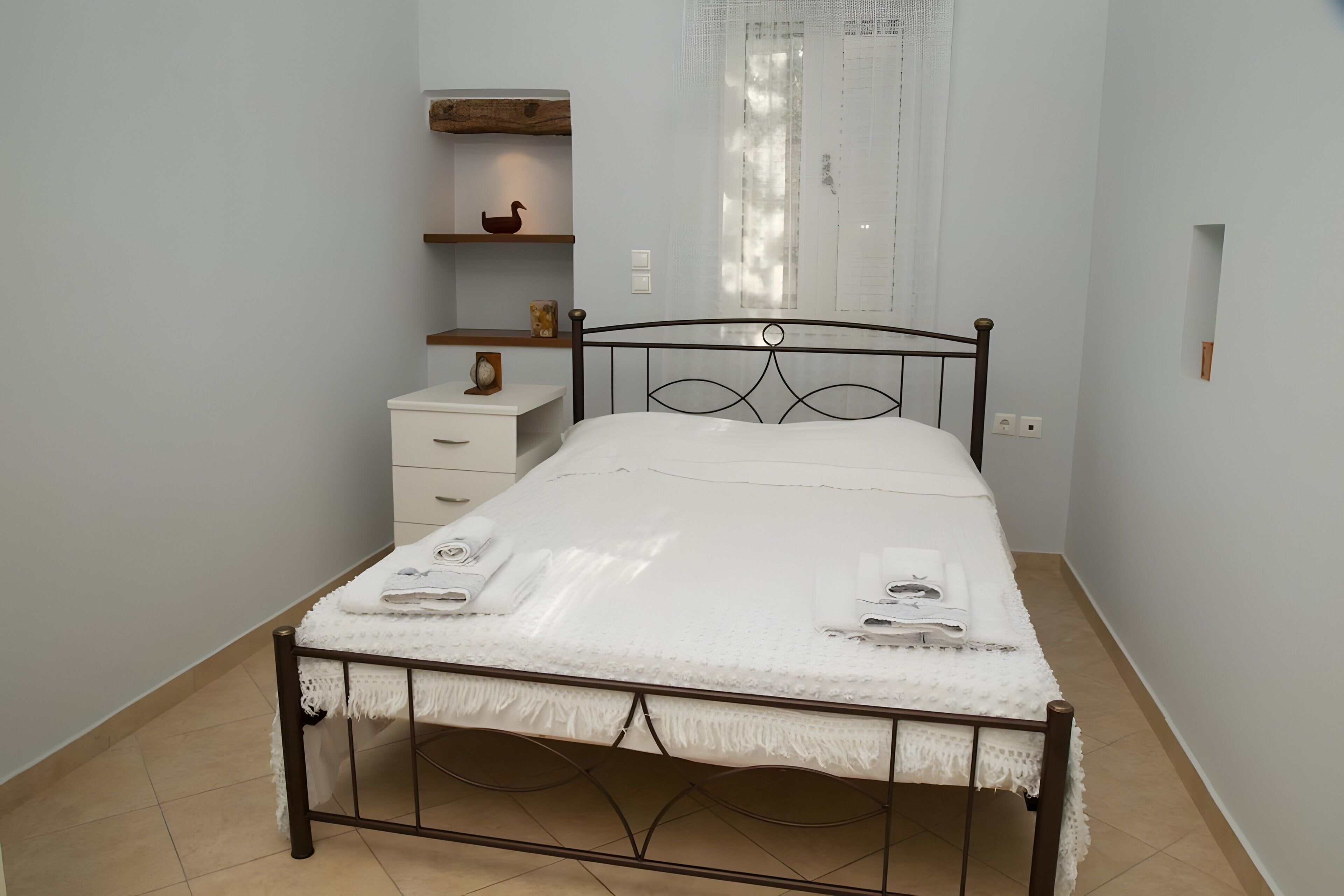 A bedroom with a single bed, a bedside table, a nightstand, a shelf with decorative items, and a window with curtains.