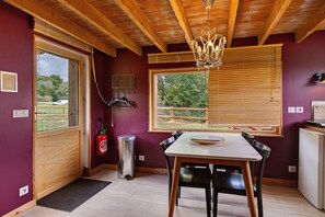 A room with a wooden ceiling, a table with chairs, a window with blinds, a fire extinguisher, and a small refrigerator. - La Petite Maison de Rose – In the Heart of the Forest (Flancourt-Crescy-en-Roumois)