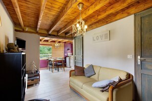 A cozy living room with a wooden ceiling, a sofa with cushions, a television, and a dining area with a table and chairs. - La Petite Maison de Rose – In the Heart of the Forest (Flancourt-Crescy-en-Roumois)