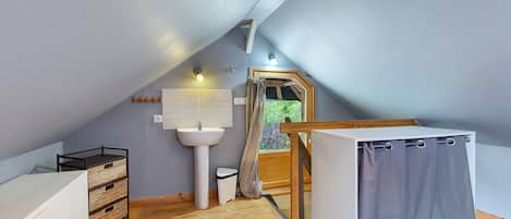 Un pequeño dormitorio en el ático con techo inclinado, suelo de madera, cómoda blanca, radiador blanco, ventana con vistas a zonas verdes y puerta de madera.
