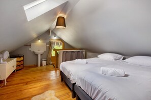A bedroom with a sloped ceiling, a bed with white linens, a bedside table with a lamp, a wooden dresser, and a window with curtains. - La Petite Maison de Rose – In the Heart of the Forest (Flancourt-Crescy-en-Roumois)