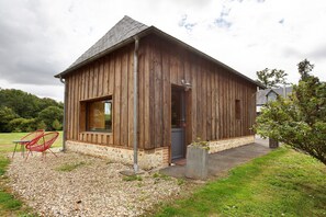 Une cabane en bois avec un toit en pente, une porte et une fenêtre. Devant la cabane, on aperçoit une chaise rouge et un pot de fleurs.