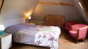 An attic bedroom with a sloped ceiling, a bed with floral bedding, a red chair, and a brick wall. - The Old Stable – In the Heart of 20 Hectares of Greenery (Flancourt-Crescy-en-Roumois)