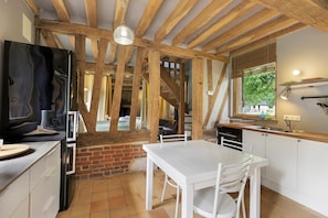 A kitchen with a white table, chairs, and a brick wall. There is a window with blinds and a hanging light fixture. - The Old Stable – In the Heart of 20 Hectares of Greenery (Flancourt-Crescy-en-Roumois)