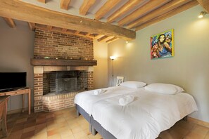 A bedroom with a brick fireplace, a bed with white linens, a painting on the wall, and a television on a wooden table. - The Old Stable – In the Heart of 20 Hectares of Greenery (Flancourt-Crescy-en-Roumois)
