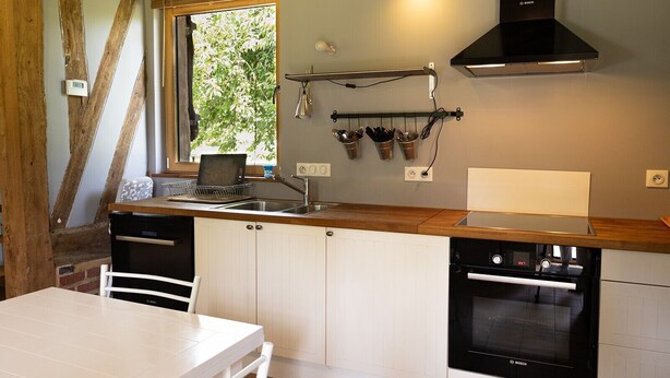 A kitchen with a Bosch oven, a window with blinds, a hanging rack with utensils, and a wooden ceiling. - The Old Stable – In the Heart of 20 Hectares of Greenery (Flancourt-Crescy-en-Roumois)