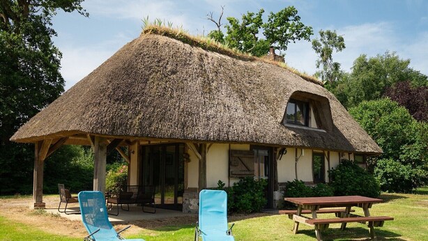 A thatched-roof house with a covered patio, two blue chairs, and a picnic table. - The Cottage – In the Heart of a 20-Hectare Estate (Flancourt-Crescy-en-Roumois)