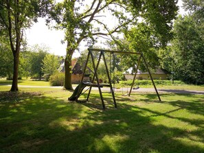 A swing set in a grassy park area with trees and a building in the background. - La Longère – In the Heart of a 20-Hectare Estate (Flancourt-Crescy-en-Roumois)