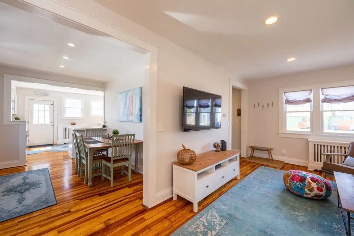 A spacious living area with a wooden floor, a dining table with chairs, a television mounted on the wall, and a colorful bean bag.