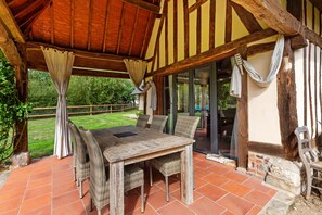 A covered patio with a wooden table and wicker chairs, a glass door leading to a garden, and a view of the outdoors. - La Roselière – In the Heart of 20 Hectares of Greenery (Flancourt-Crescy-en-Roumois)