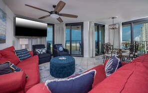 A modern living room with a flat-screen TV, red and blue furniture, a ceiling fan, and a balcony with a view of the city. - Phoenix Vii 7501 3 Bedroom Condo (Orange Beach)
