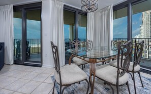 A dining area with a glass table and chairs, a view of the ocean through large windows, and a balcony with a view of the cityscape. - Phoenix Vii 7501 3 Bedroom Condo (Orange Beach)