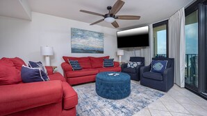 Ein modernes Wohnzimmer mit einem roten Sofa, einem blauen Hocker und dunklen Sesseln, einem Deckenventilator, einem Flachbildfernseher und einem großen Fenster mit Vorhängen.
