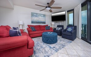 A modern living room with a red sofa, blue ottoman, and dark armchairs, a ceiling fan, a flat-screen TV, and a large window with curtains. - Phoenix Vii 7501 3 Bedroom Condo (Orange Beach)