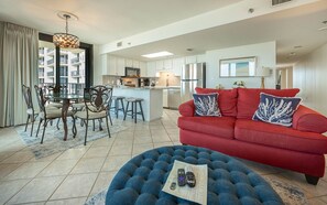 A modern living room with a red sofa, a blue ottoman, and a dining area with a glass table and chairs. - Phoenix Vii 7501 3 Bedroom Condo (Orange Beach)