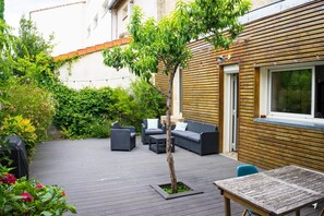 Eine Terrasse im Freien mit Holzdeck, einem kleinen Baum und Gartenmöbeln, darunter eine Couch und Stühle.