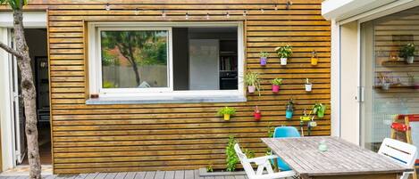 A modern outdoor patio with a wooden deck, a table with chairs, and a window overlooking a garden.
