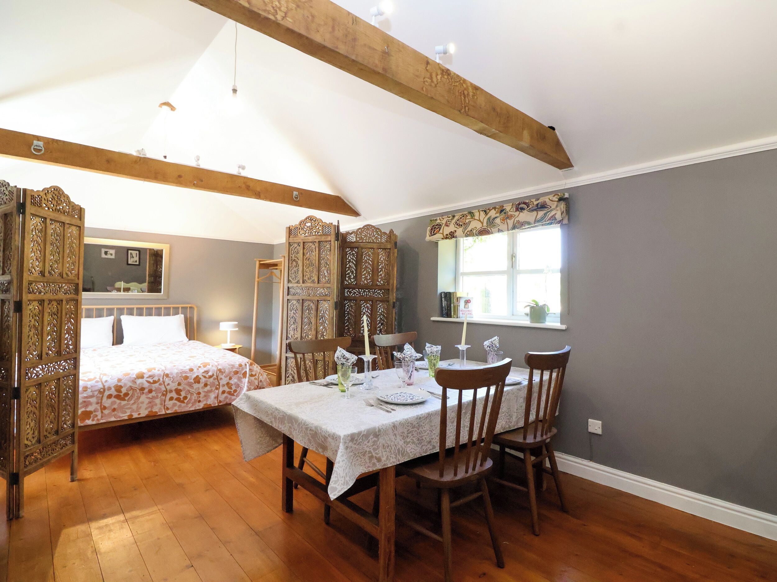 Una habitación con una mesa de comedor preparada para cuatro personas, sillas de madera, una cama con colcha estampada y una ventana con vistas.