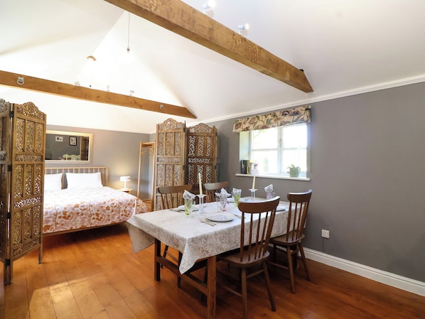 A room with a dining table set for four, wooden chairs, a bed with a floral bedspread, and a window with a view.