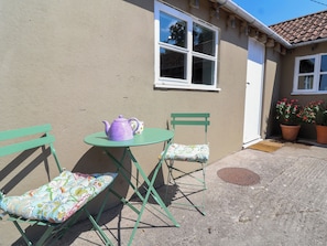 A small outdoor seating area with a table and two chairs, a window, and potted plants.