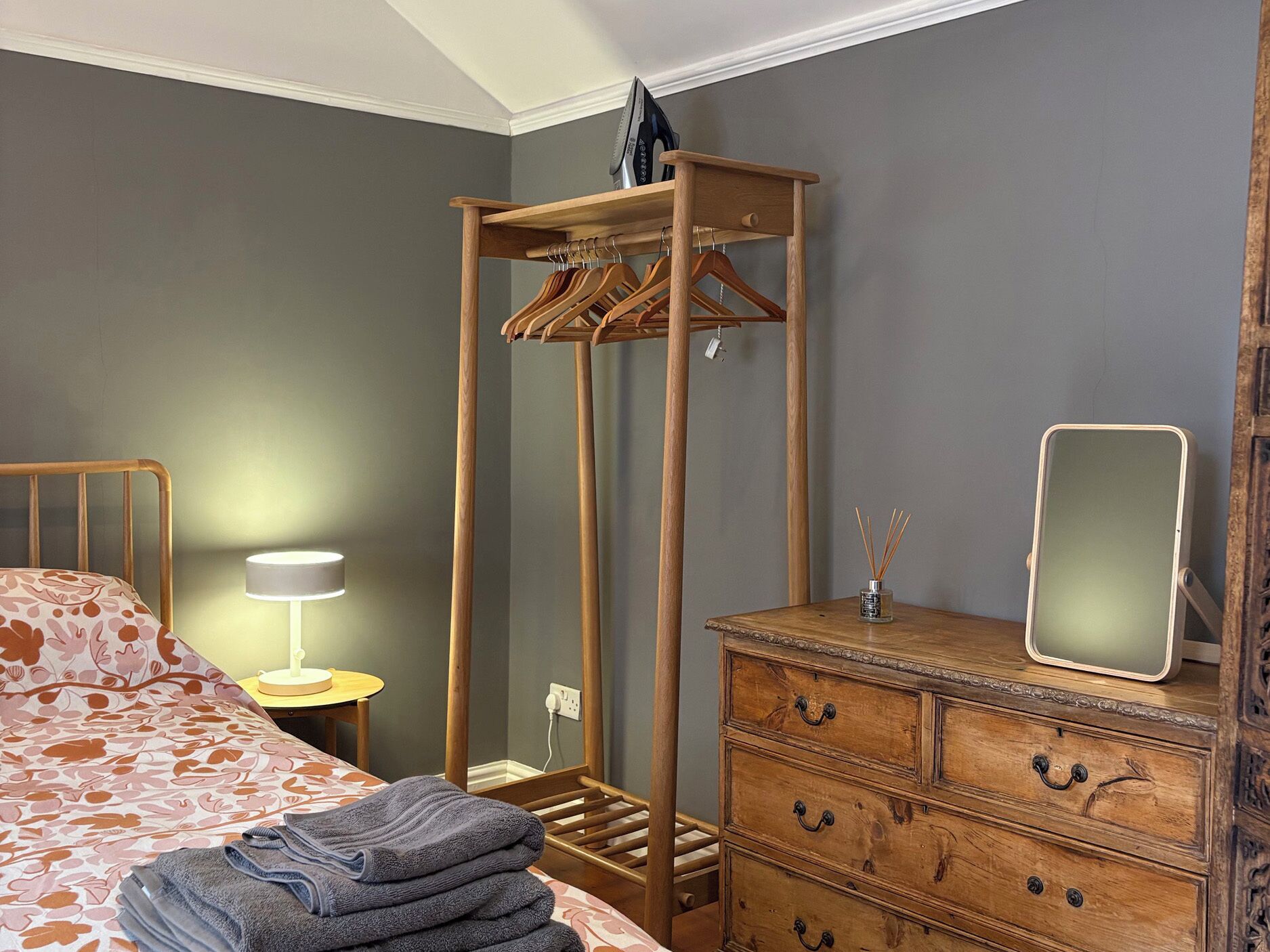 A bedroom with a bed, a wooden dresser, a mirror, a hanging clothes rack, and a bedside lamp.