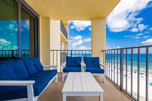 Un balcon avec une table blanche et des chaises bleues, donnant sur une plage parsemée de parasols.