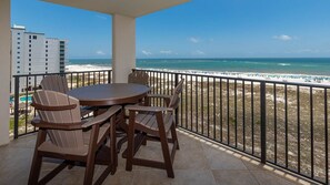 A balcony with a table and chairs, overlooking a beach with a clear view of the ocean and a distant building. - Phoenix Vii 7710 2 Bedroom Condo (Orange Beach)
