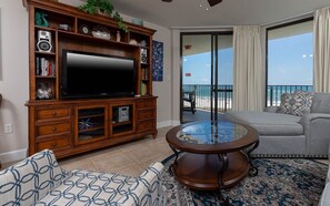 A living room with a flat-screen TV, wooden entertainment unit, patterned sofa, and a glass coffee table. - Phoenix Vii 7710 2 Bedroom Condo (Orange Beach)