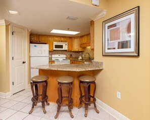 Une cuisine avec des meubles en bois, un réfrigérateur blanc, un four à micro-ondes, une cuisinière et un plan de travail en granit. On y trouve trois tabourets de bar et un tableau encadré accroché au mur.