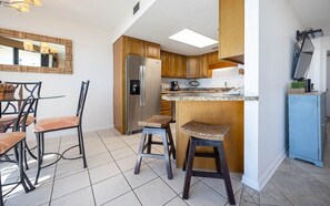 A modern kitchen with a refrigerator, wooden cabinets, a bar stool, and a dining table with chairs. - Phoenix Vii 7314 1 Bedroom Condo (Orange Beach)