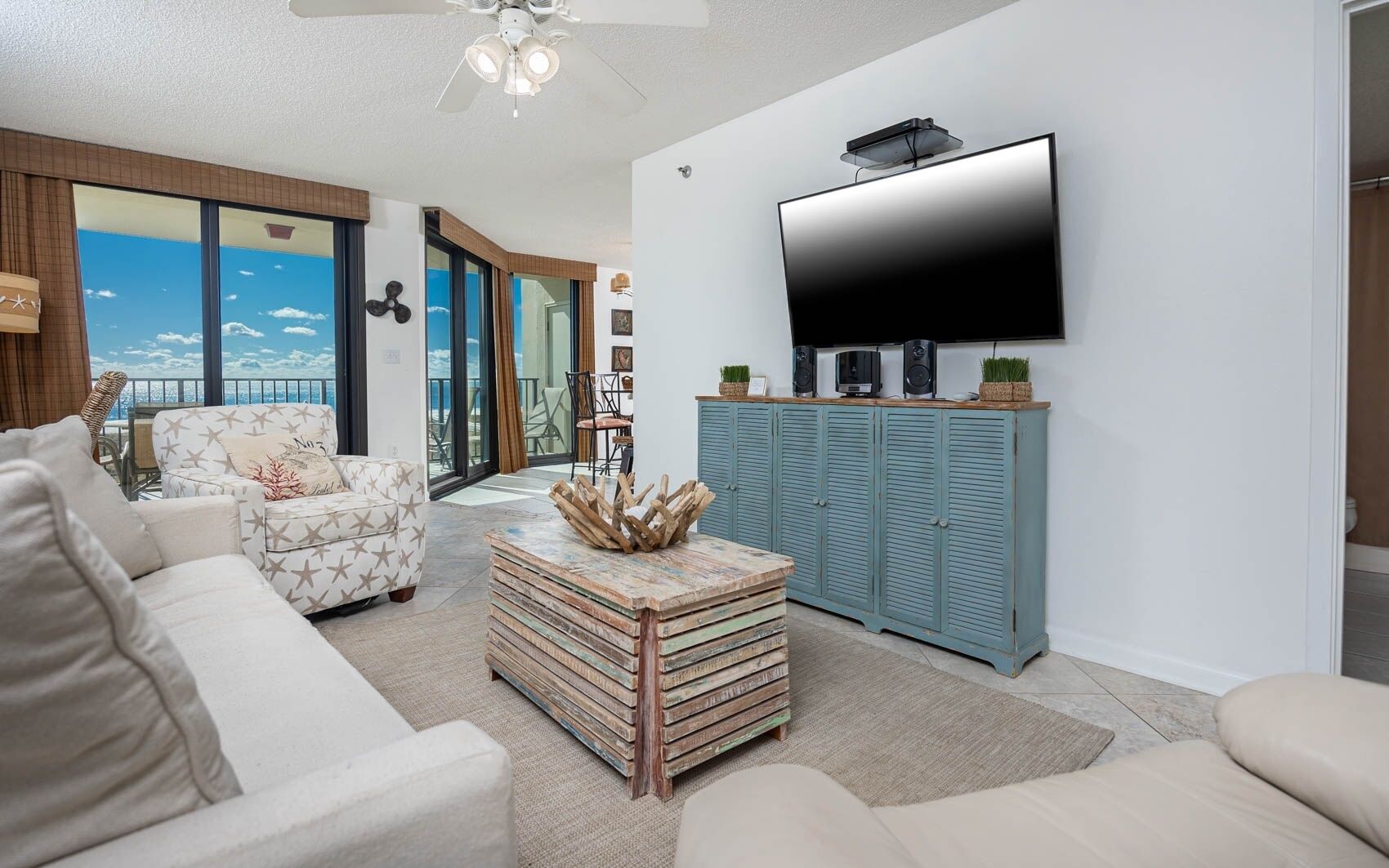 Een woonkamer met een grote flatscreen televisie, een plafondventilator en uitzicht op de oceaan door grote ramen.