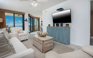 A living room with a large flat-screen TV, a ceiling fan, and a view of the ocean through large windows. - Phoenix Vii 7314 1 Bedroom Condo (Orange Beach)