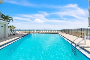 Ein rechteckiger Swimmingpool mit klarem, blauem Wasser, umgeben von einem schwarzen Metallzaun, und eine Reihe weißer Liegestühle auf der linken Seite.