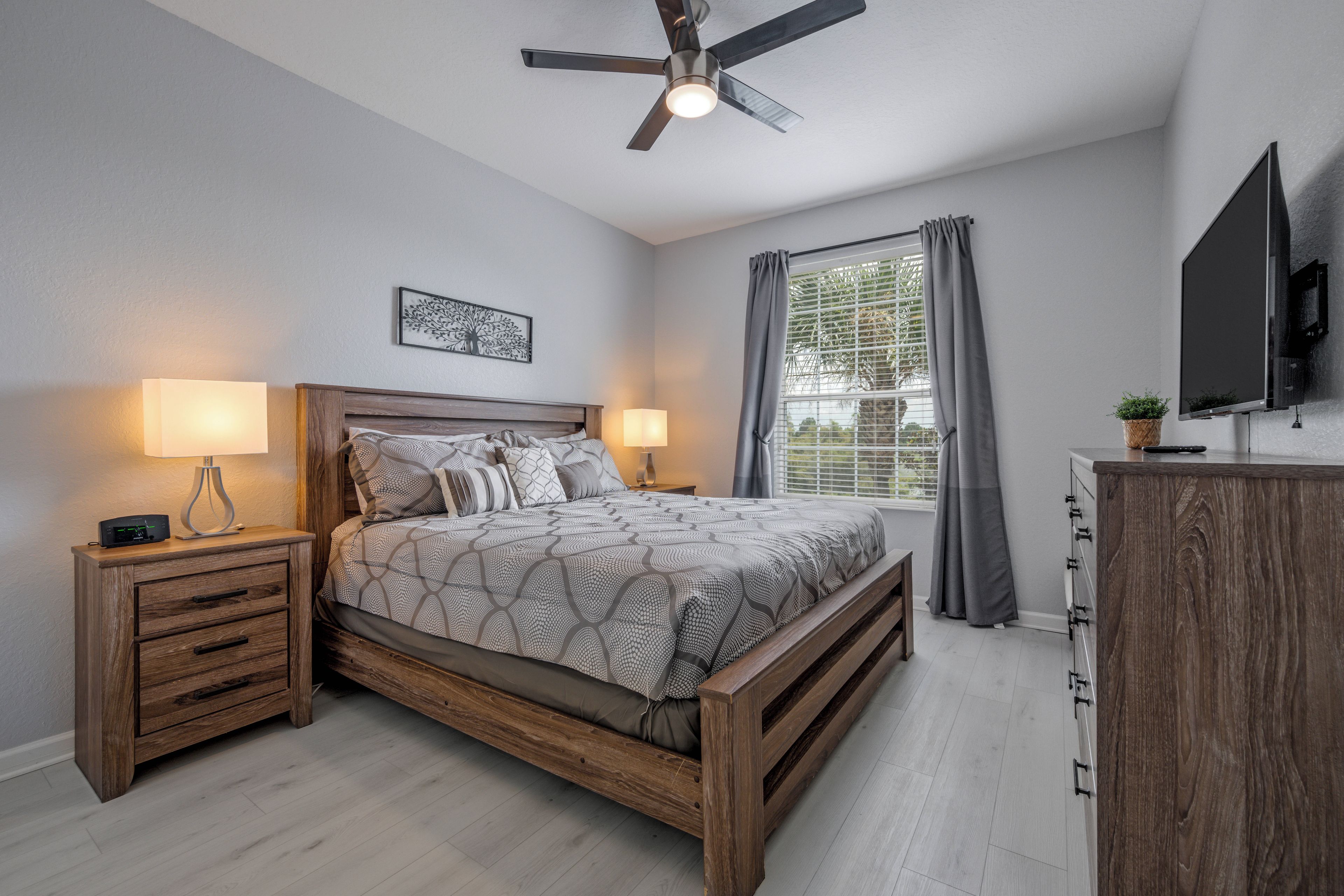 Une chambre avec un lit en bois, deux tables de chevet avec des lampes, un ventilateur de plafond, un téléviseur fixé au mur et une fenêtre avec des rideaux.